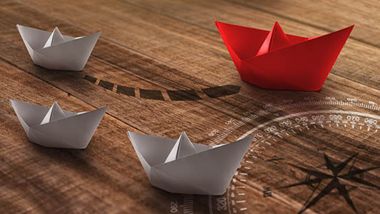 Business or travel concept: a group of white paper ships heading in one direction and one red paper ship individual pointing in the different way as a business,, travel or shipping industry icon for innovative solution.